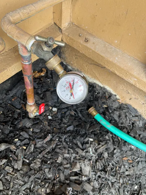 A brass pressure gauge attached to an outdoor hose bib reading about 100 PSI during a water pressure test